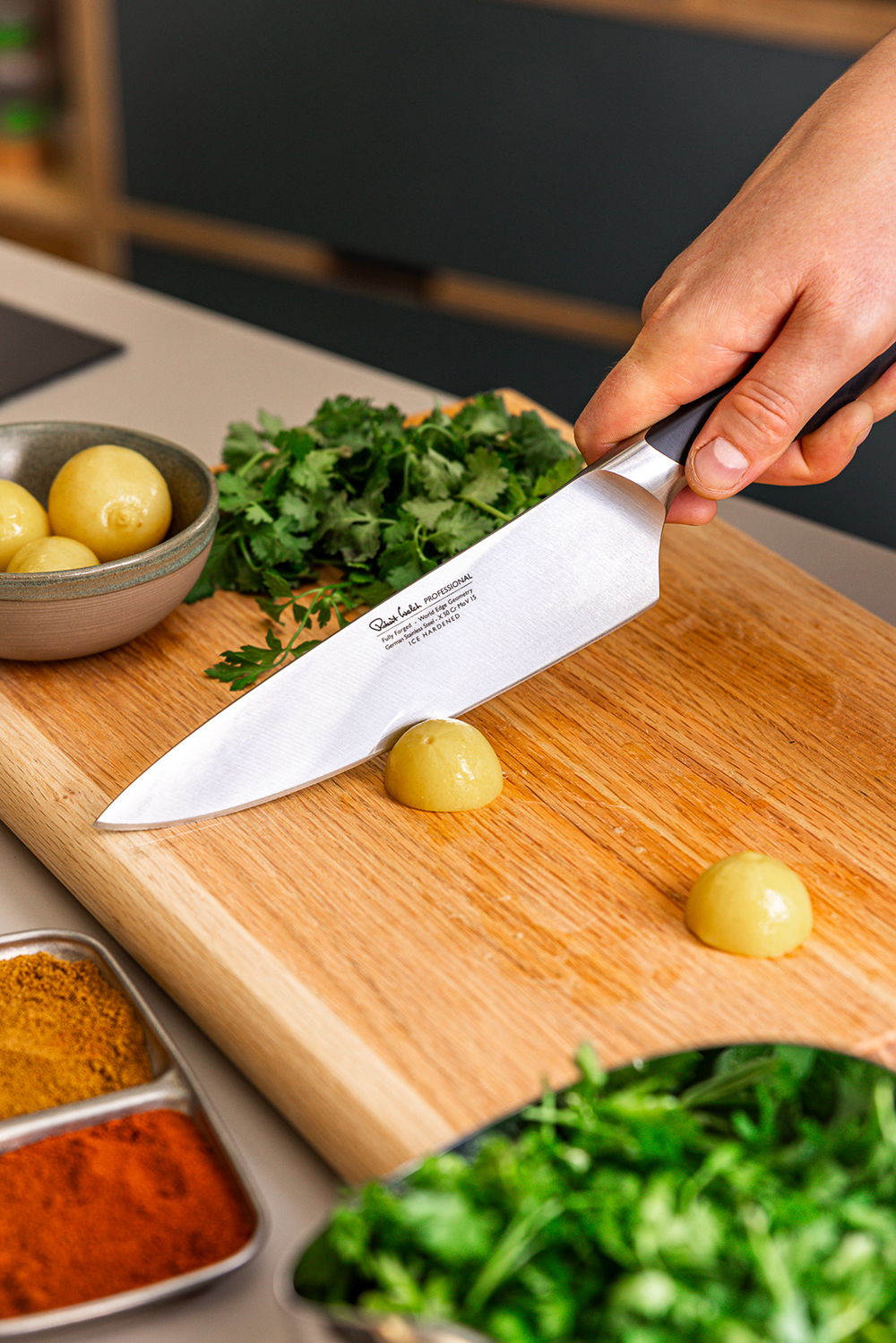 A hand holding a Professional Chef's Knife above small potatoes on a Classic Chopping Board.