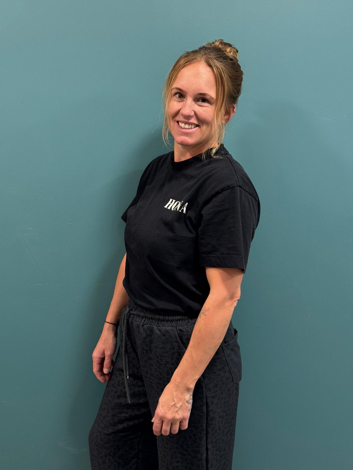 Smiling woman in black shirt and gray pants against a teal background.