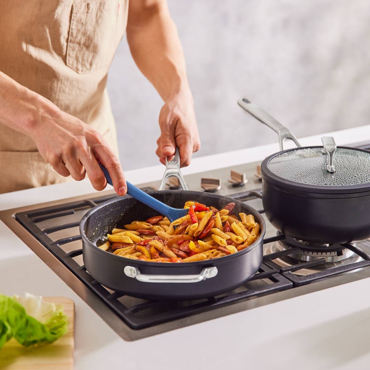A chef’s hand uses a Misen Silicone Spoontula to stir pasta in a Misen Nonstick Sauté on a stovetop, next to a Misen Nonstick Saucier with a lid on.