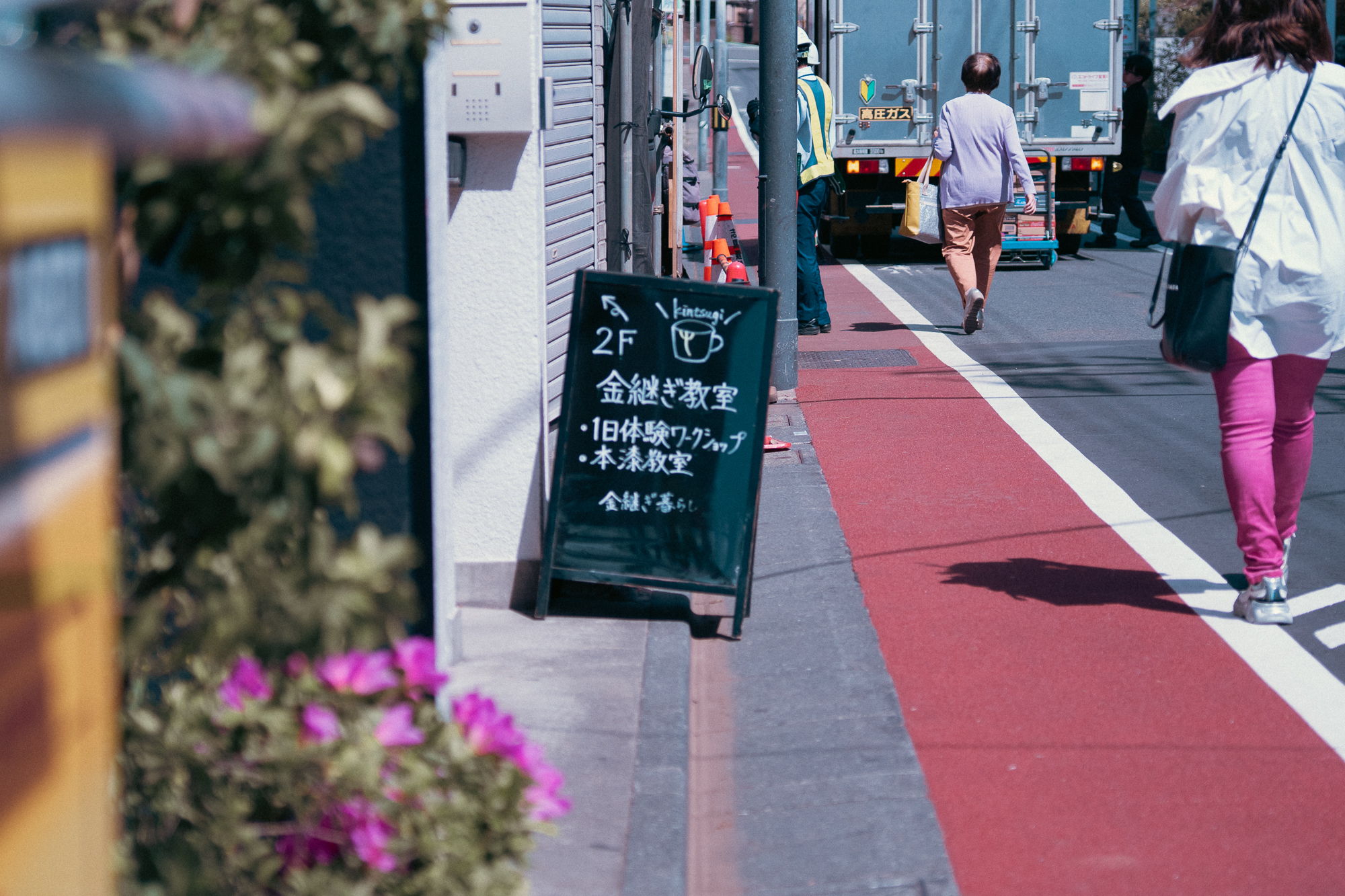 Sidewalk with a sign, flowers, and pedestrians walking in the background.
