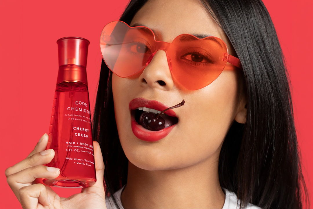 Woman wearing red sunglasses holding a beauty product against a pink background.