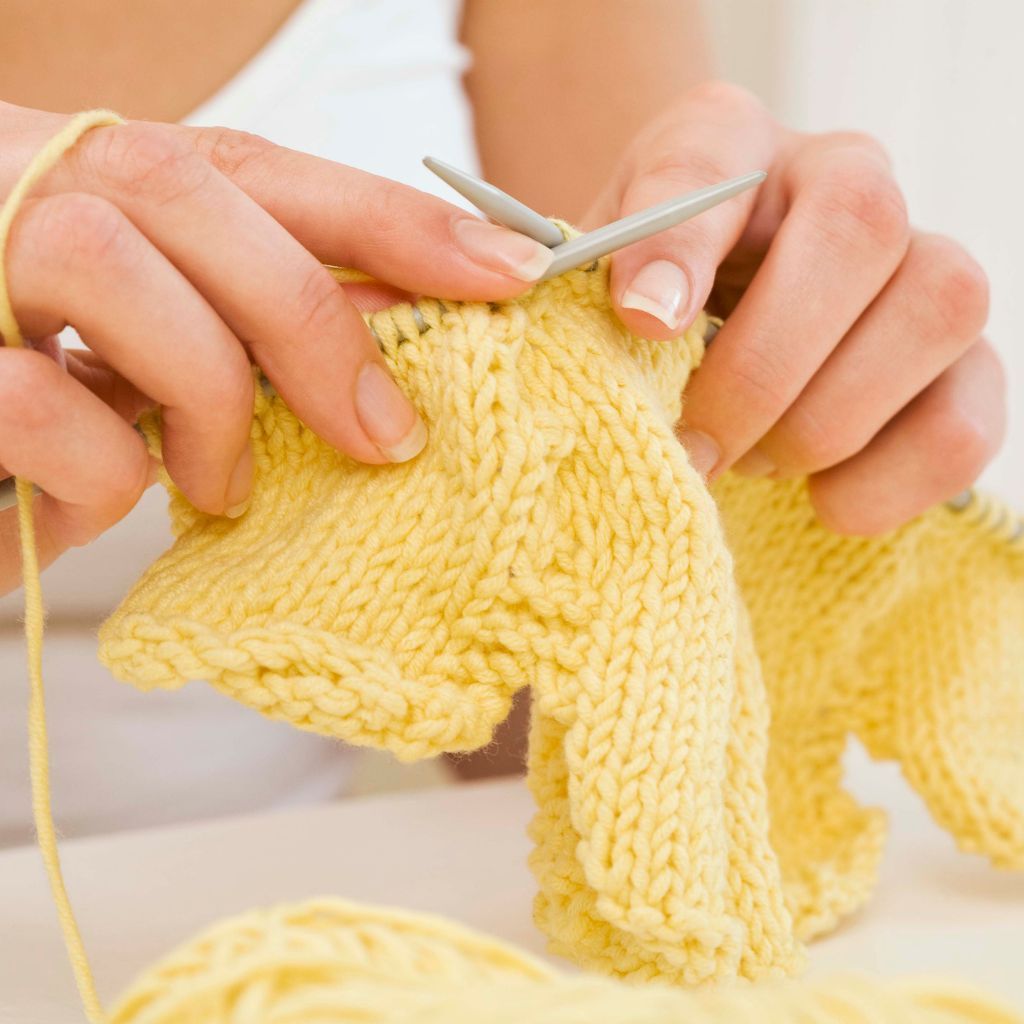 Person die ein gelbes Kleidungsstück strickt.