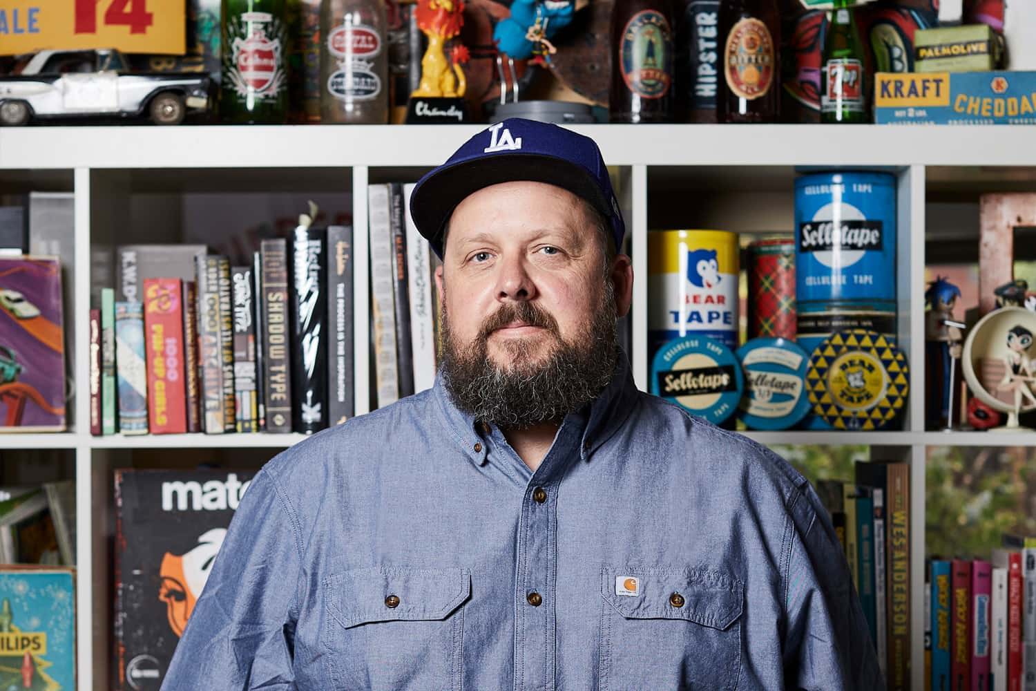A photograph of Travis Price in front of a wall of books and ephemera.
