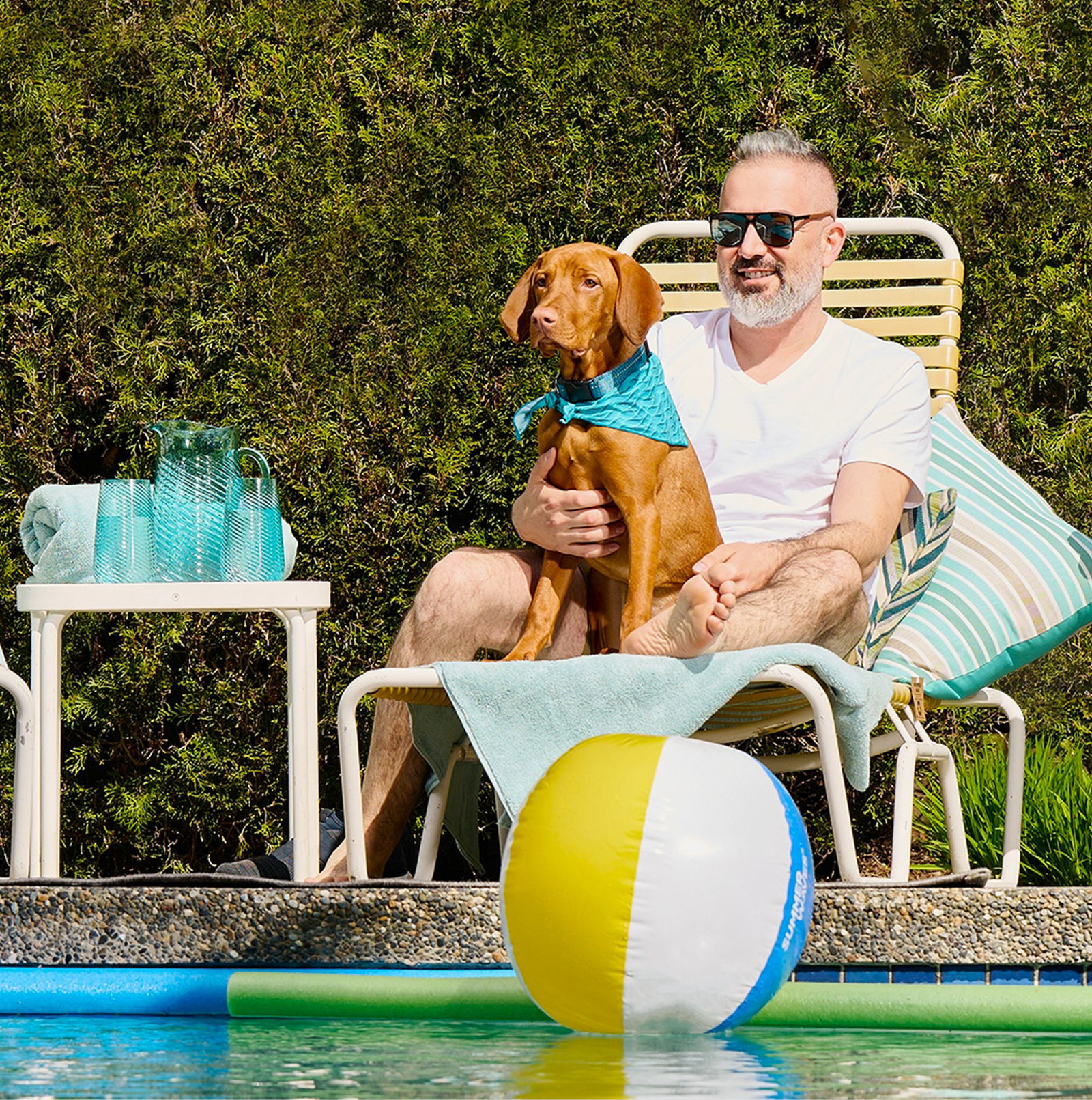 Man relaxing by pool with dog and beach ball.