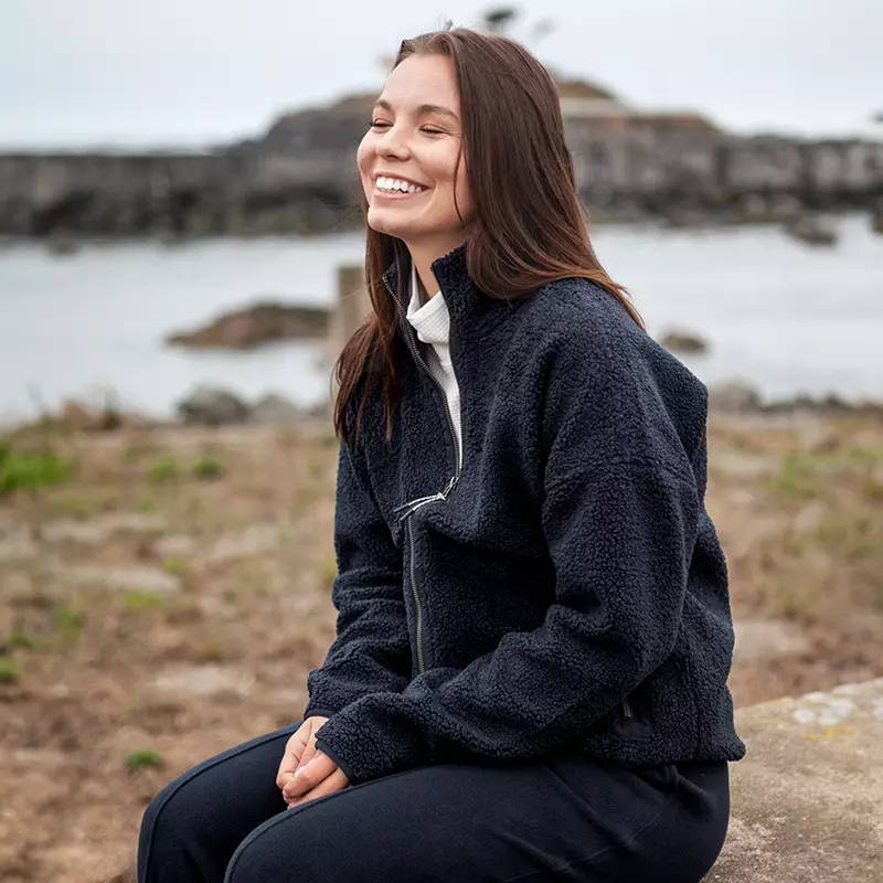 Woman sitting on rock at beach in recycled polyester pullover.