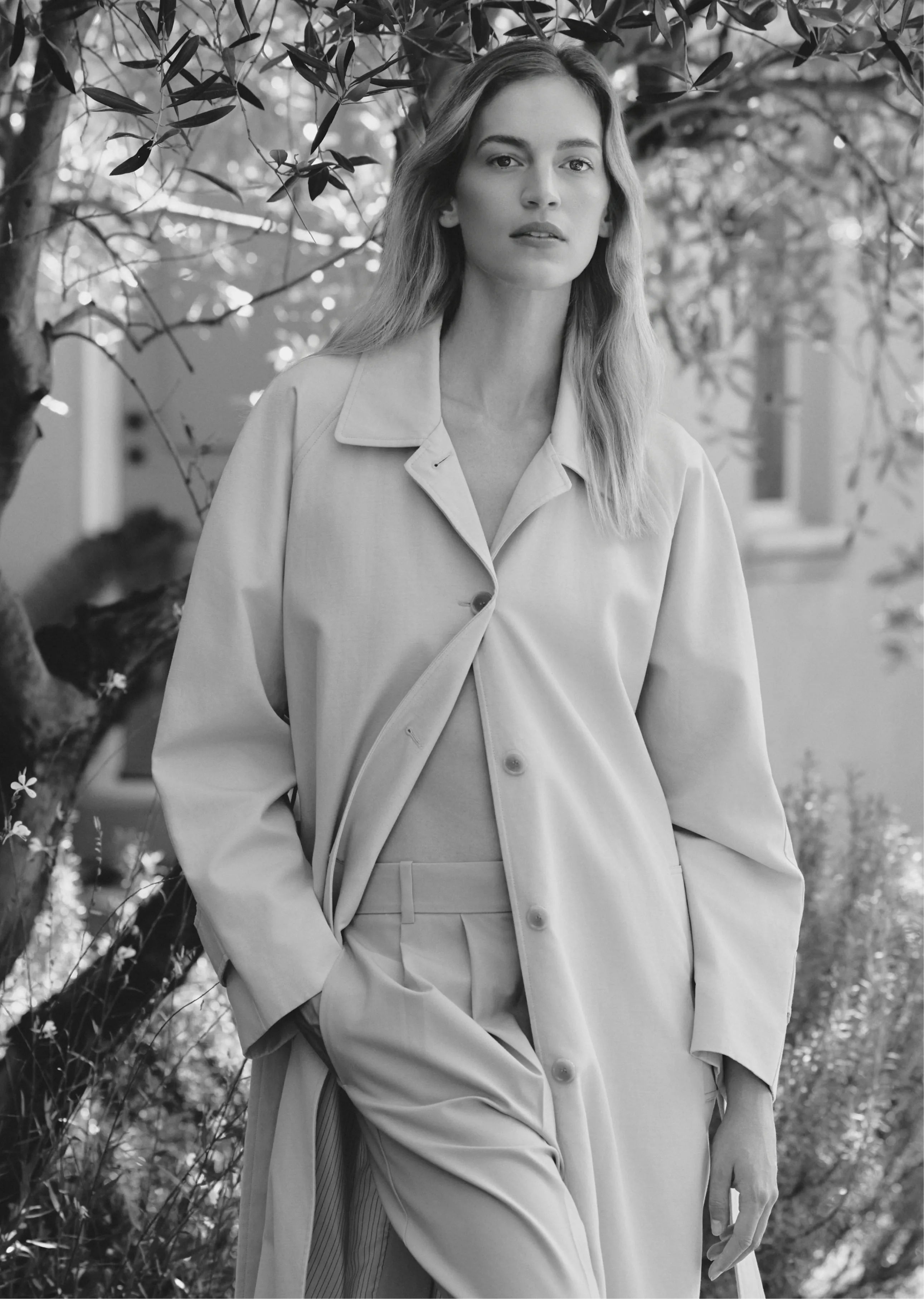 A woman in a light coat stands outdoors among greenery.