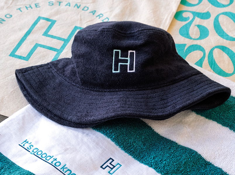 Navy terry towel bucket hat with embroidered H logo displayed on top of a folded striped teal and white beach towel and screenprinted tote bag