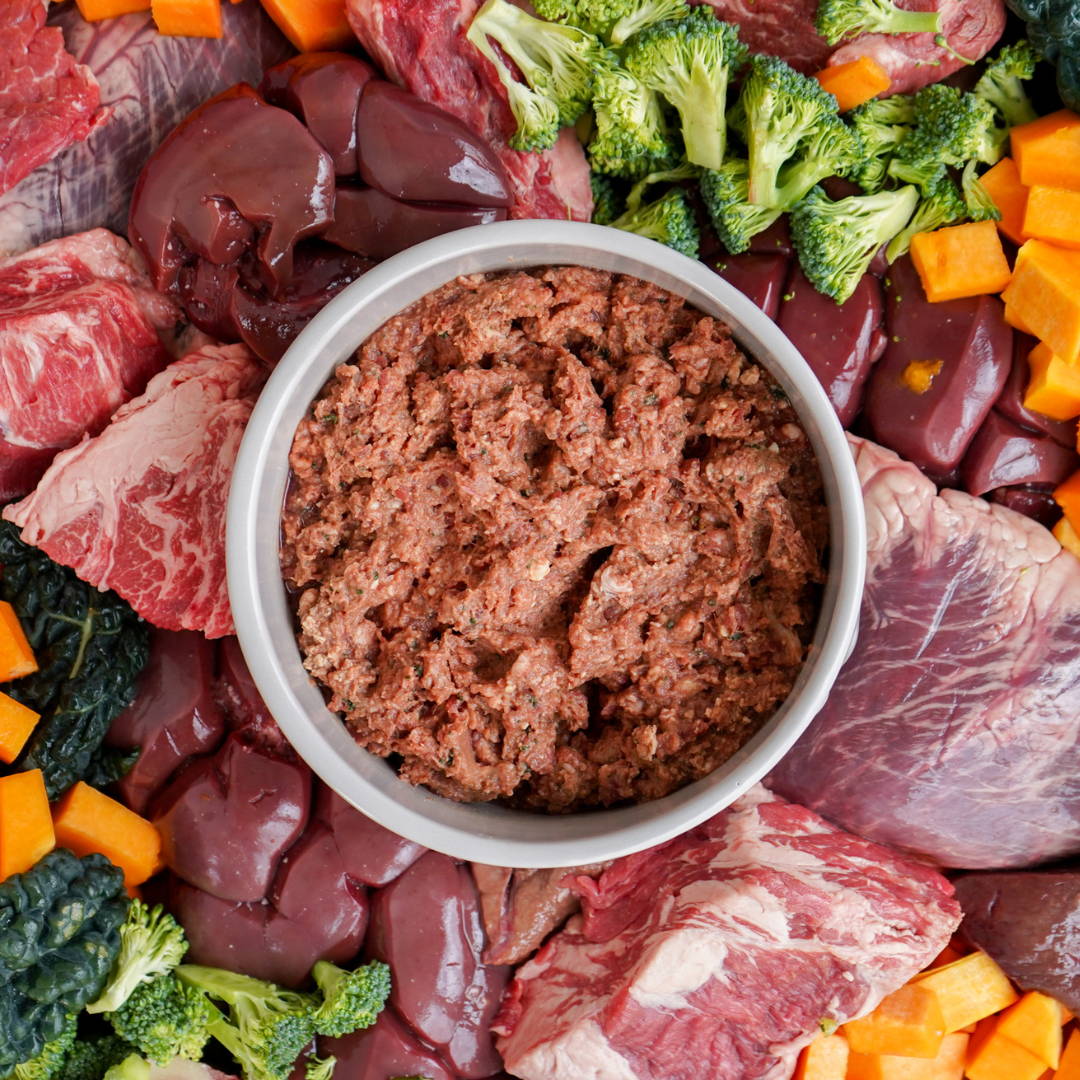White bowl filled with ground raw dog food surrounded by raw vegetables and meat.