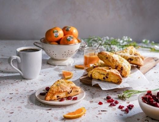 Coffee, scones, and oranges on a table with flowers and fruit.