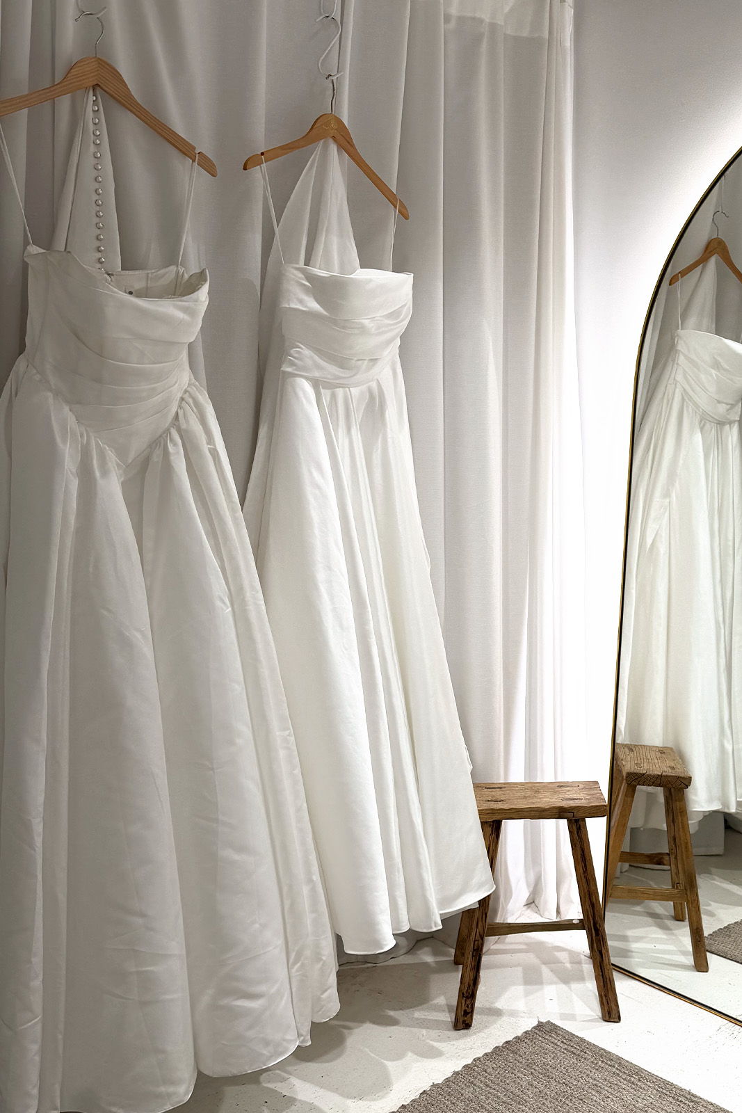 Two elegant white dresses hanging in a fitting room with a mirror.