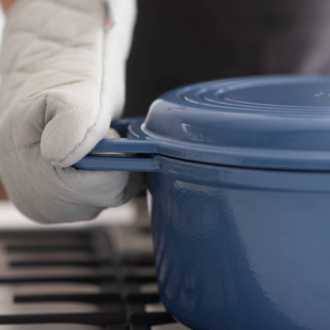 Hand with gray oven mitt firmly gripping the handles of a Blue Misen Dutch Oven with Grill Lid.