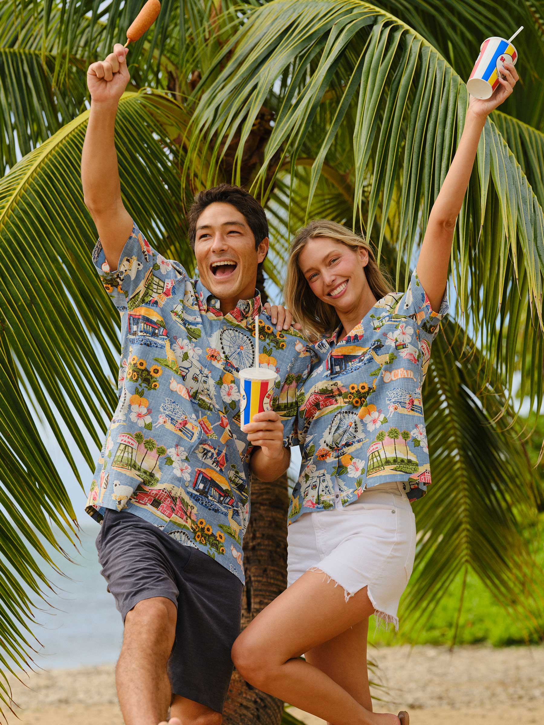 Two people in tropical shirts celebrating by a palm tree at the beach.