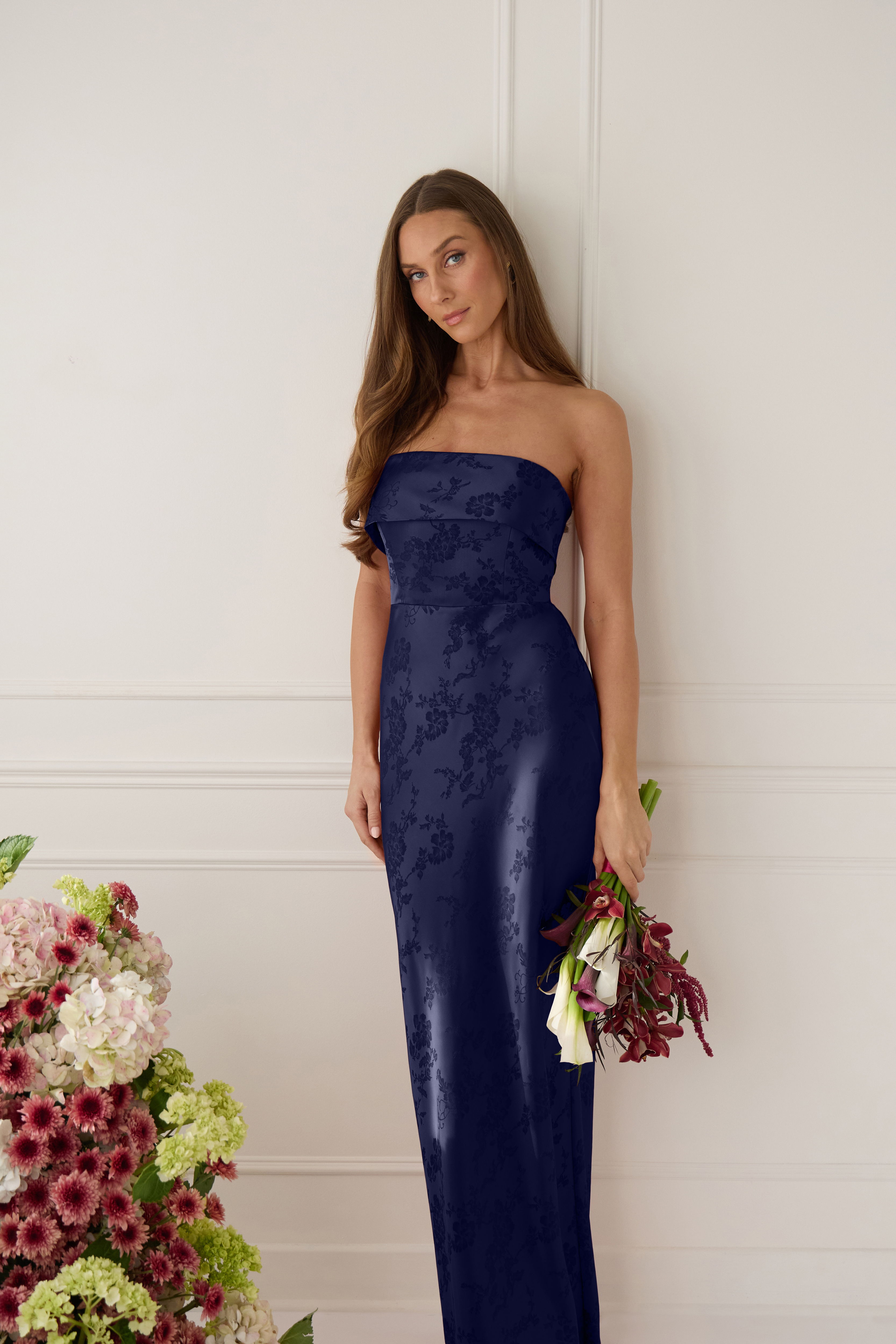 Woman in a strapless navy gown holding flowers, standing near floral arrangement.