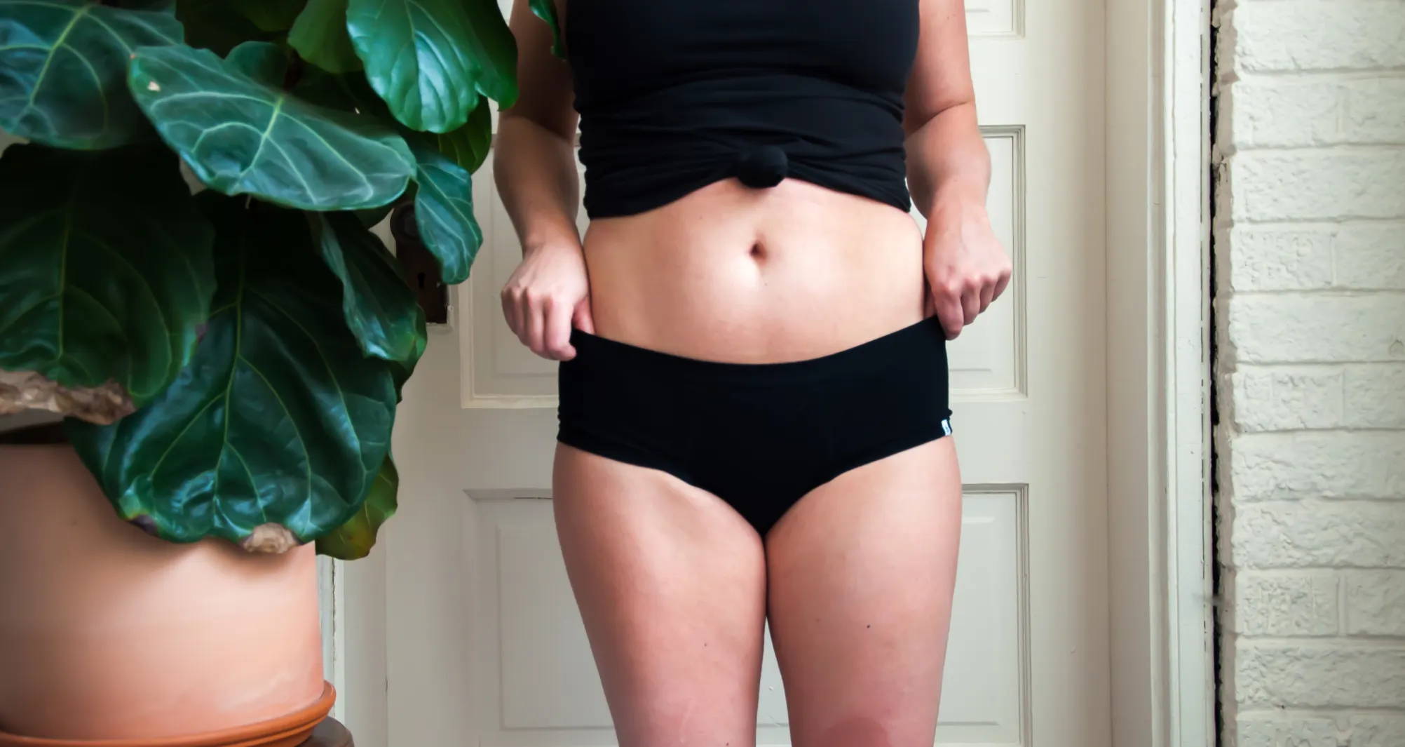 A woman wearing a black shirt and black underwear stands in front of a white door and next to a large leafy plant.