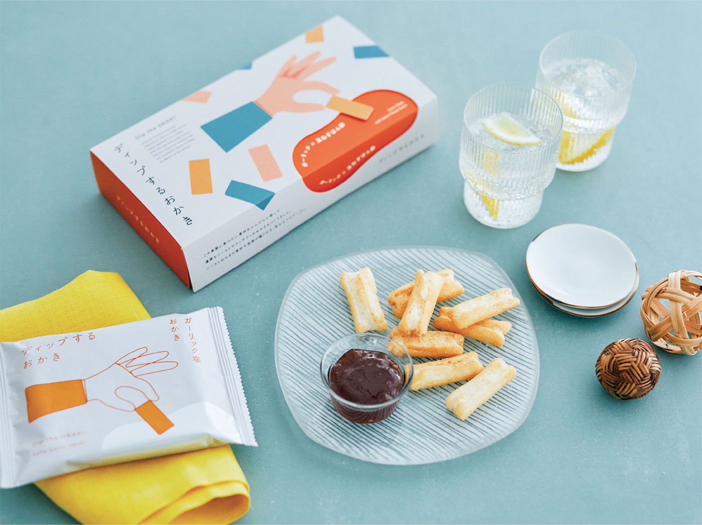 A bowl of dip surrounded by snacks and an orange on a yellow background.