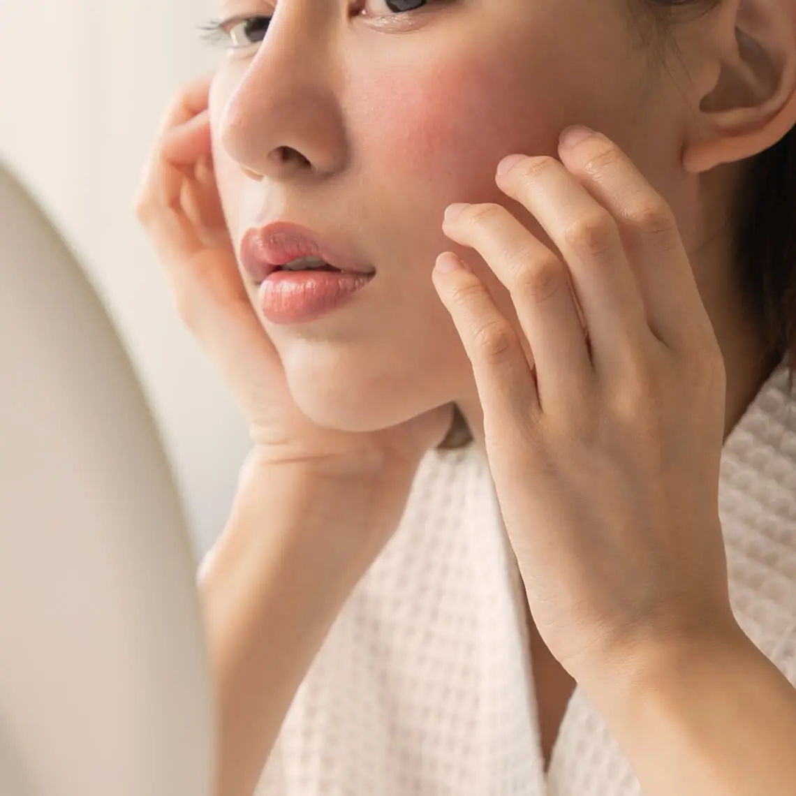 Woman examining her face in a mirror, touching her cheeks.