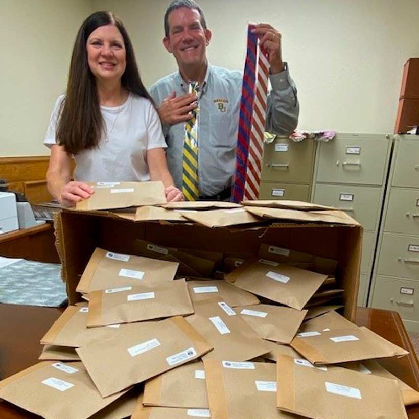 A man and women holding up ties and standing in front of a box of packaged ties waiting to be mailed