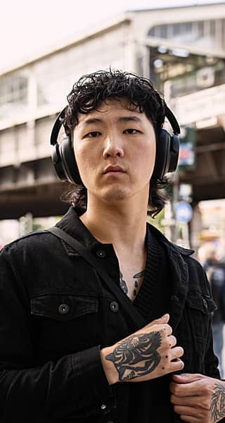 Young individual wearing headphones, standing outdoors with urban background.