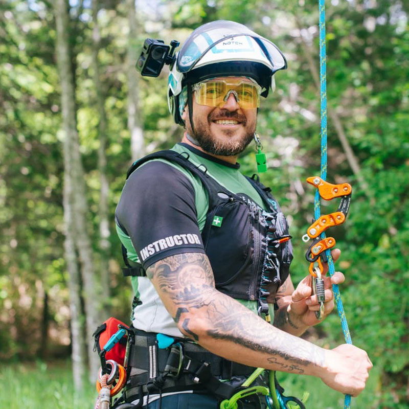Man in climbing gear smiling in a forest setting.
