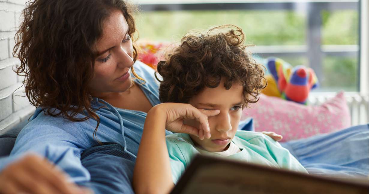 Tired boy rubbing his itchy eye while he sits on the sofa with his mother and a book – his allergies are acting up
