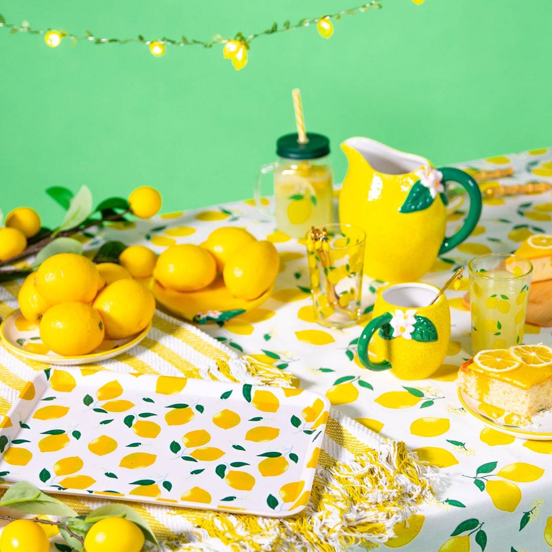 Eine lebhafte Sommertischdekoration mit einer Zitronenkollektion, darunter ein Zitronenkrug, Gläser mit Zitronenscheiben und eine Tischdecke mit Zitronenmotiven vor einem grünen Hintergrund.