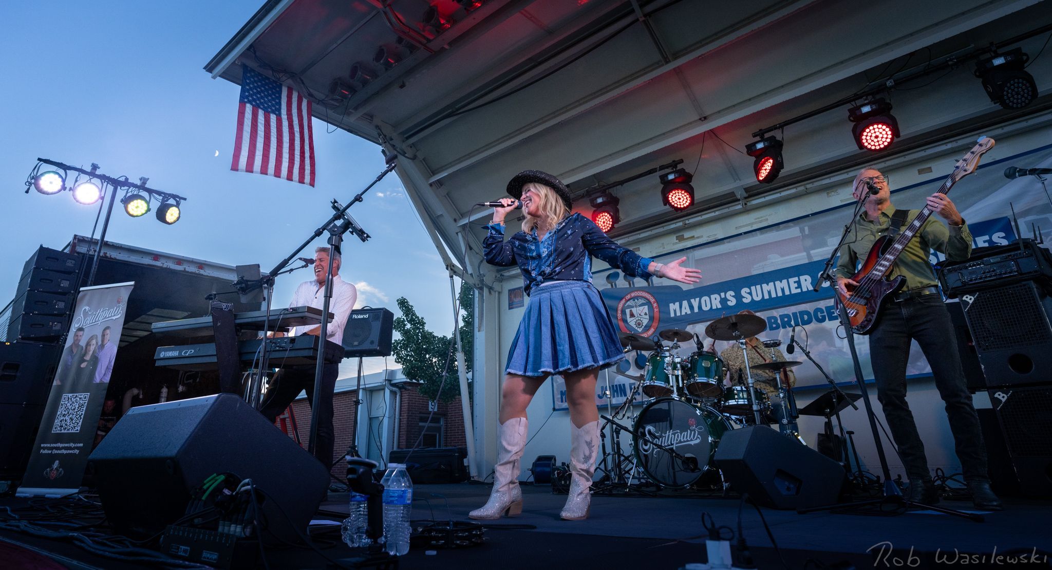 Woman singing on stage with musicians and lights, American flag in background.