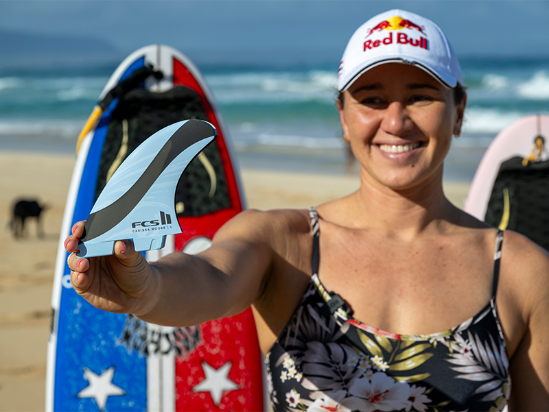 Smiling woman holding a surfboard fin on the beach.