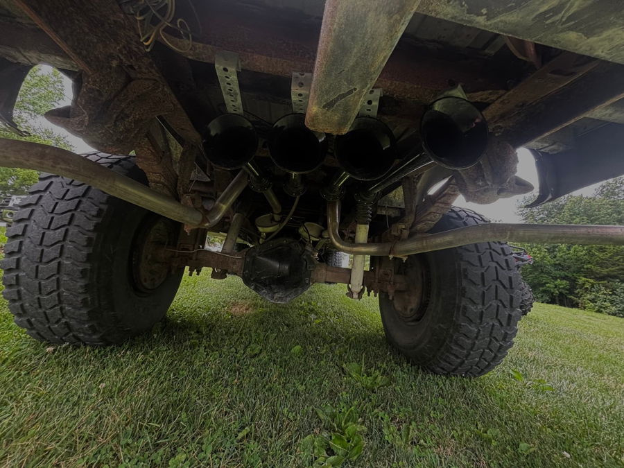 Low-angle rear underbody view four black HornBlasters Conductor's Special 127H train horn trumpets mounted below the bed facing rear with air lines rear axle dual exhaust and off-road tires Jacob's 1986 Chevy K30 on grass customer install gallery photo 2