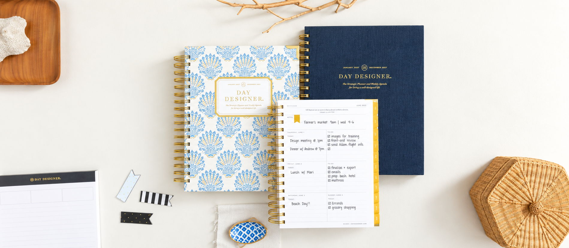 stacked 2027 day designer planners showing weekly layout, navy blue, blue and gold shelled pattern, beige background, notepad with black trim, wicker basket, tray with coral, bookmark clips