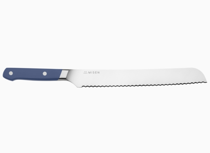 A blue Misen Serrated Knife resting on a light wood cutting board, with a sliced loaf of rustic bread visible in the background.