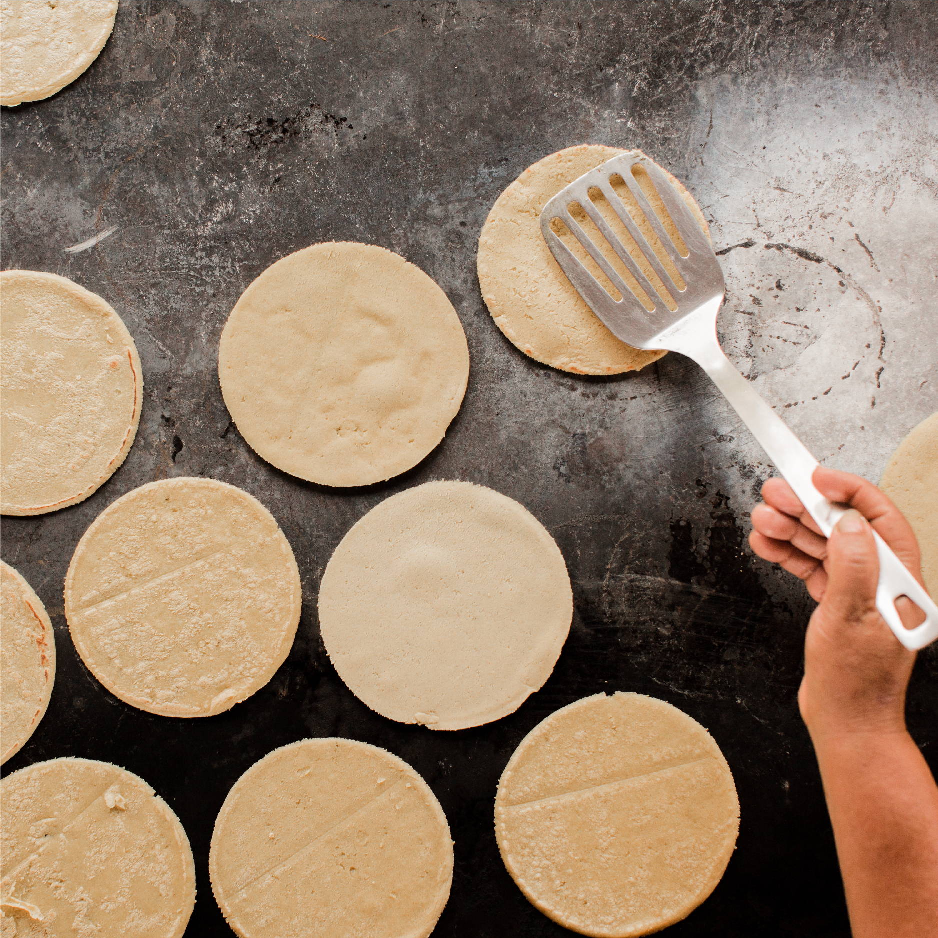 Fresh corn tortillas are cooked on a wood-fired cast iron skillet and being flipped by a spatula. 