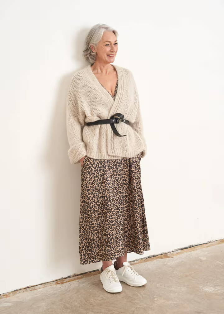 A model wearing a off white chunky knit cardigan with a black belt oveer a leopard print dress and white trainers