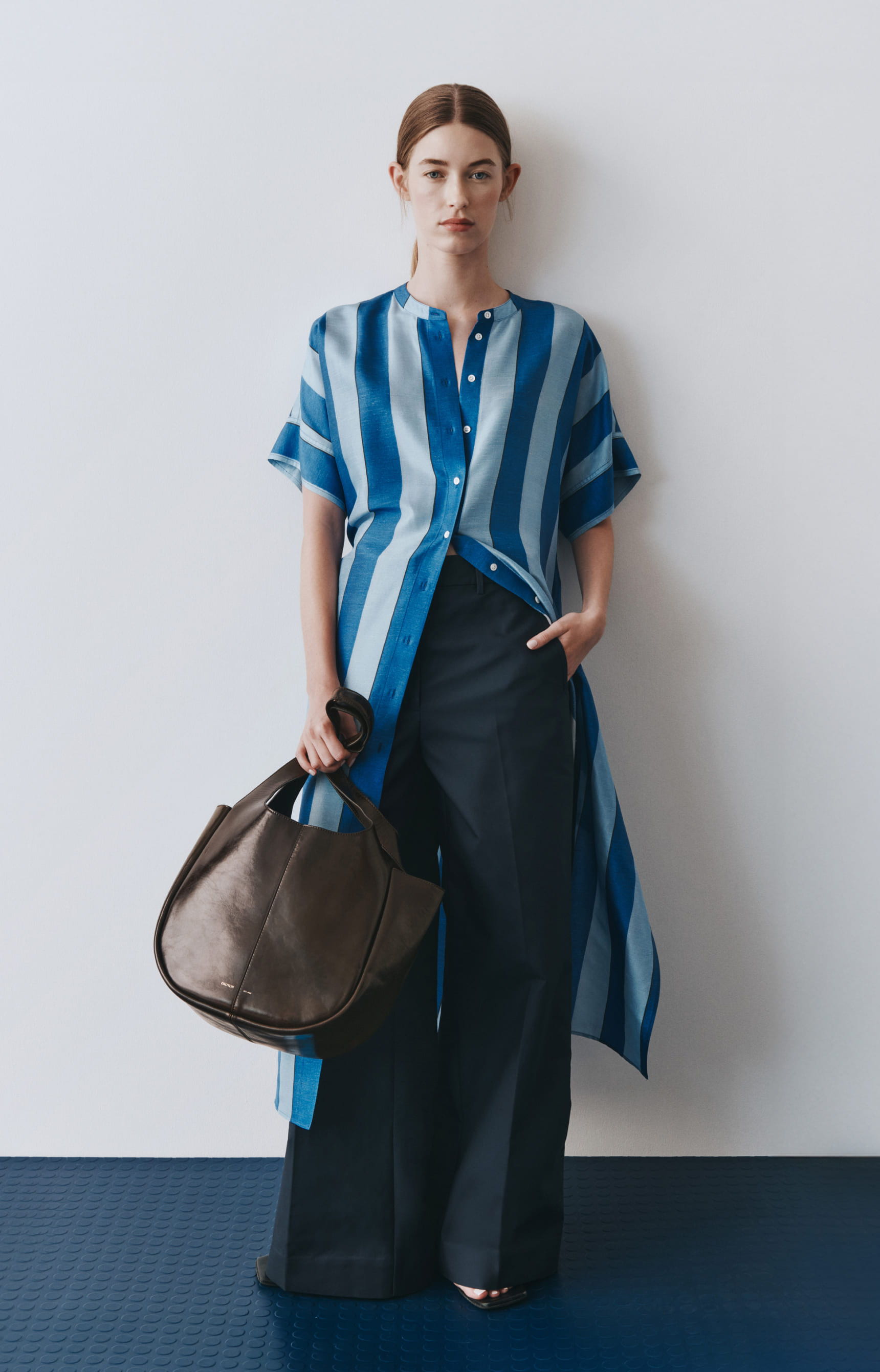 Model in blue striped shirt and dark pants, holding a handbag.