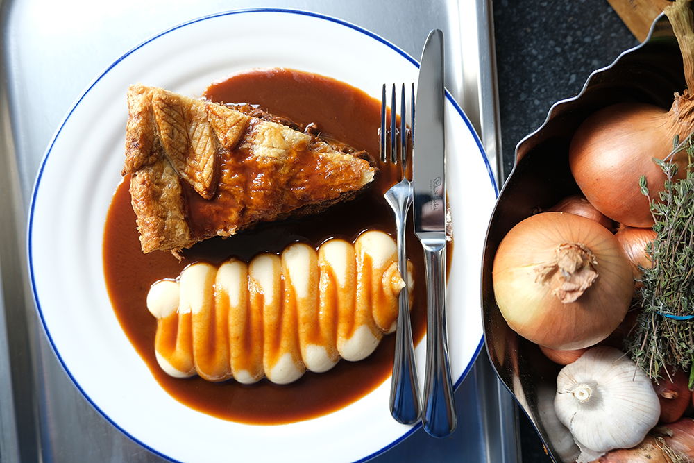 A plate with a lamb shank pie, sauce, and mashed potatoes beside onions and herbs.