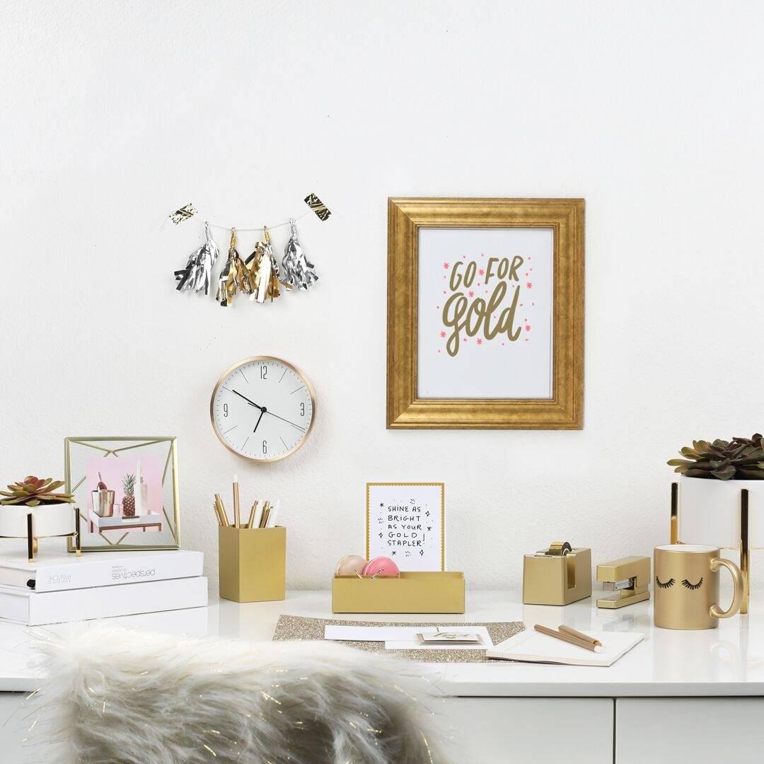 a white desk with a gold desk set, gold mug, a gold picture frame, and metallic tassels on the wall