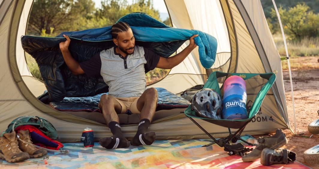 A man is sitting in a tent with his gear, throwing a Rumpl blanket around his shoulders