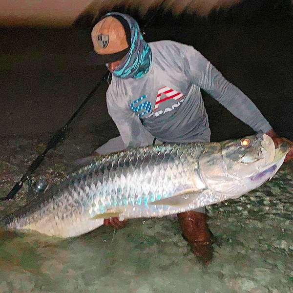 Borris Moore holding onto a very large fish on the beach, while wearing a SA Company shirt, a face shield and a SA Company hat.
