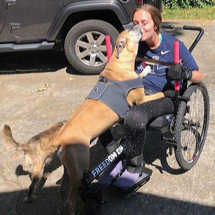 Woman holds levers of GRIT Freedom Chair off road wheelchair outdoors and kisses dog on hind legs and greeting her in her lap