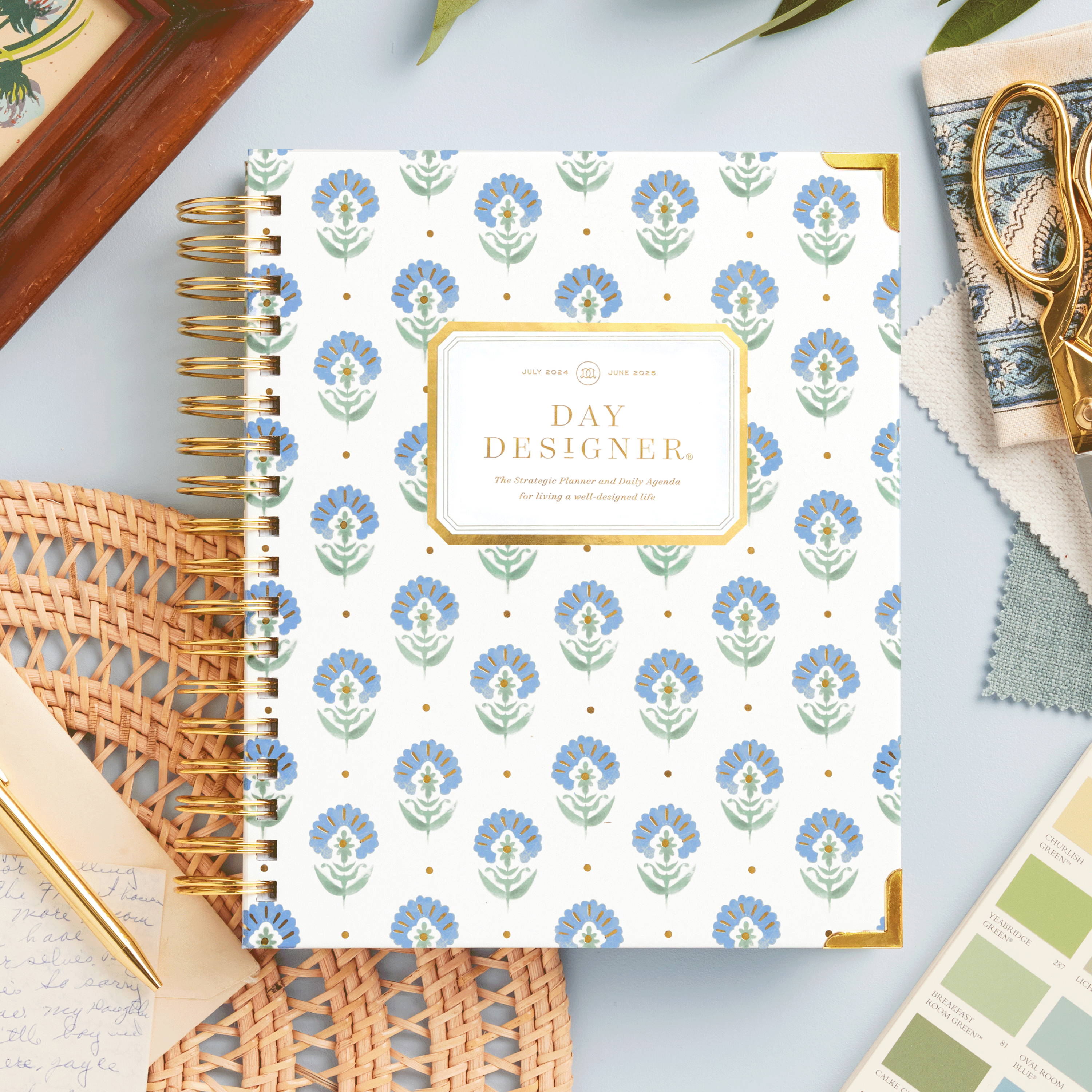 blue and green flower pattern closed book planner on light blue background, with wood picture frame, wicker placement, gold pen and stationery, paint color swatch and fabric, gold scissors