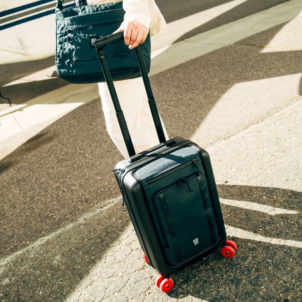 A person pulling a black suitcase with red wheels and carrying a bag.