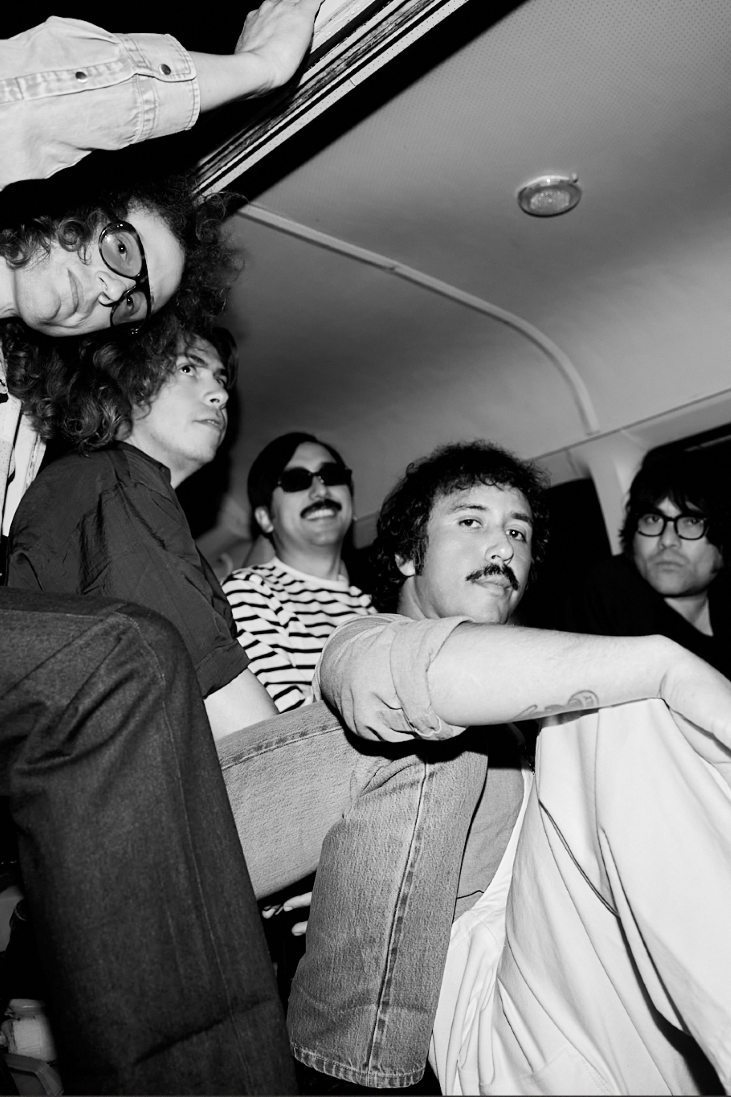Five men with varying hairstyles sitting together in a vehicle.