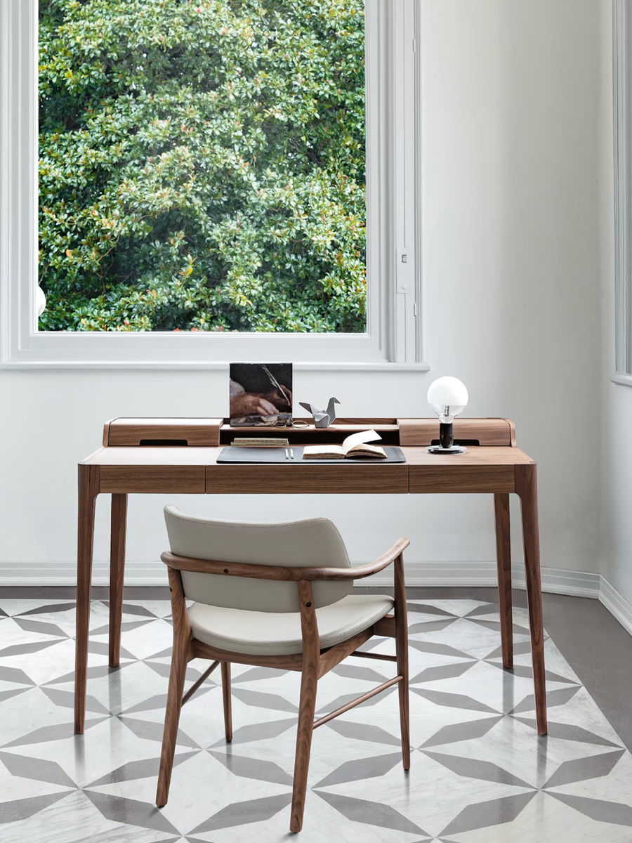 A modern desk with a chair, lamp, and window view of greenery.