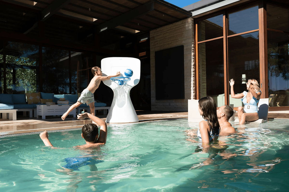 Family playing pool basketball with an in-pool hoop in a backyard pool, with an outdoor seating area under a covered patio behind.
