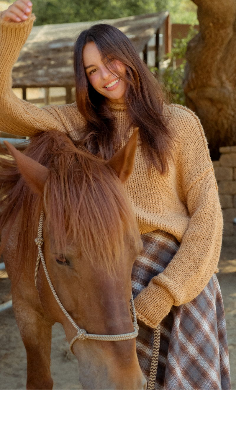 Smiling person in a sweater sitting on a horse in a sunny outdoor setting.