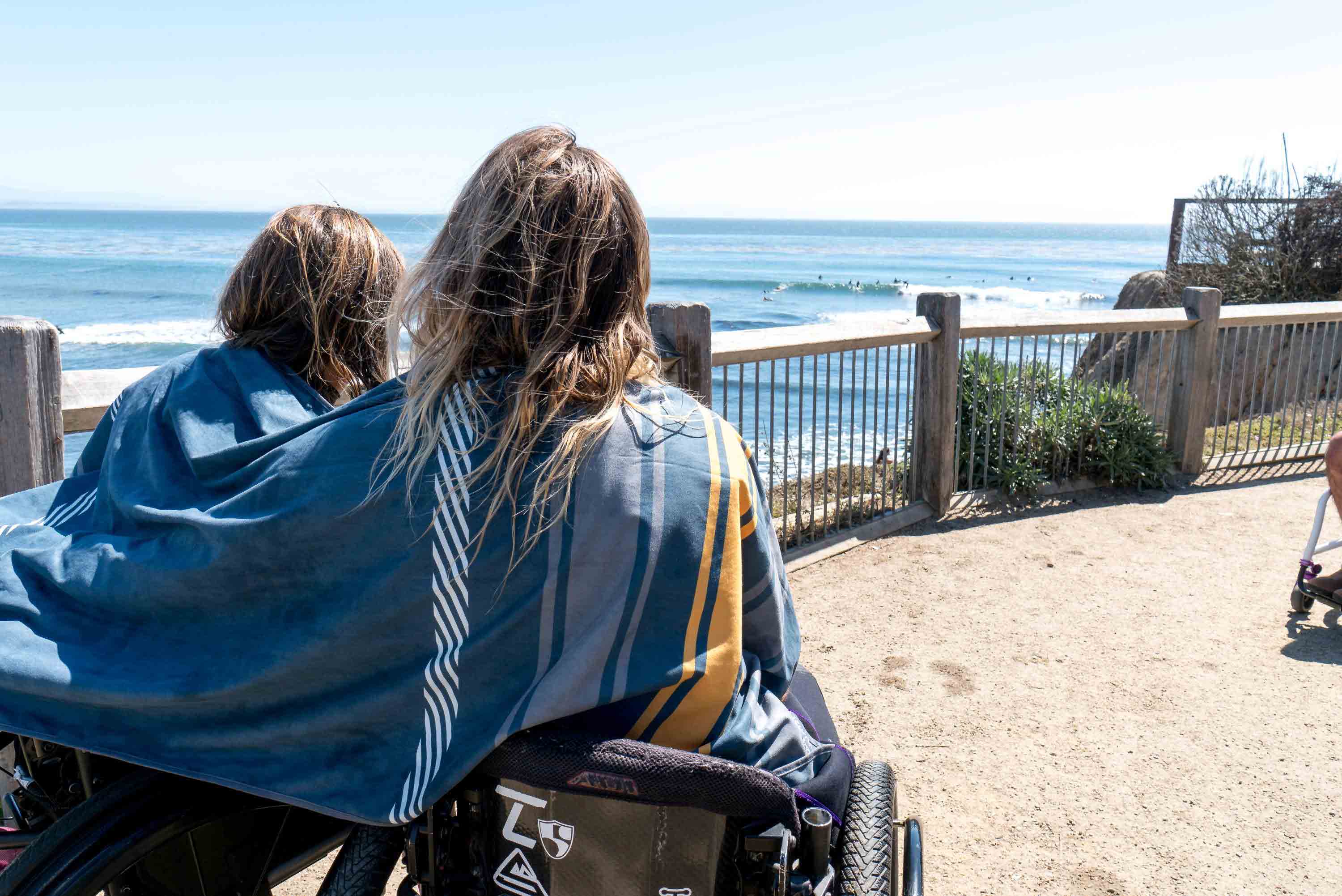 A man and women sitting in wheel chairs with High Fives Shammy wrapped around them watching surfers