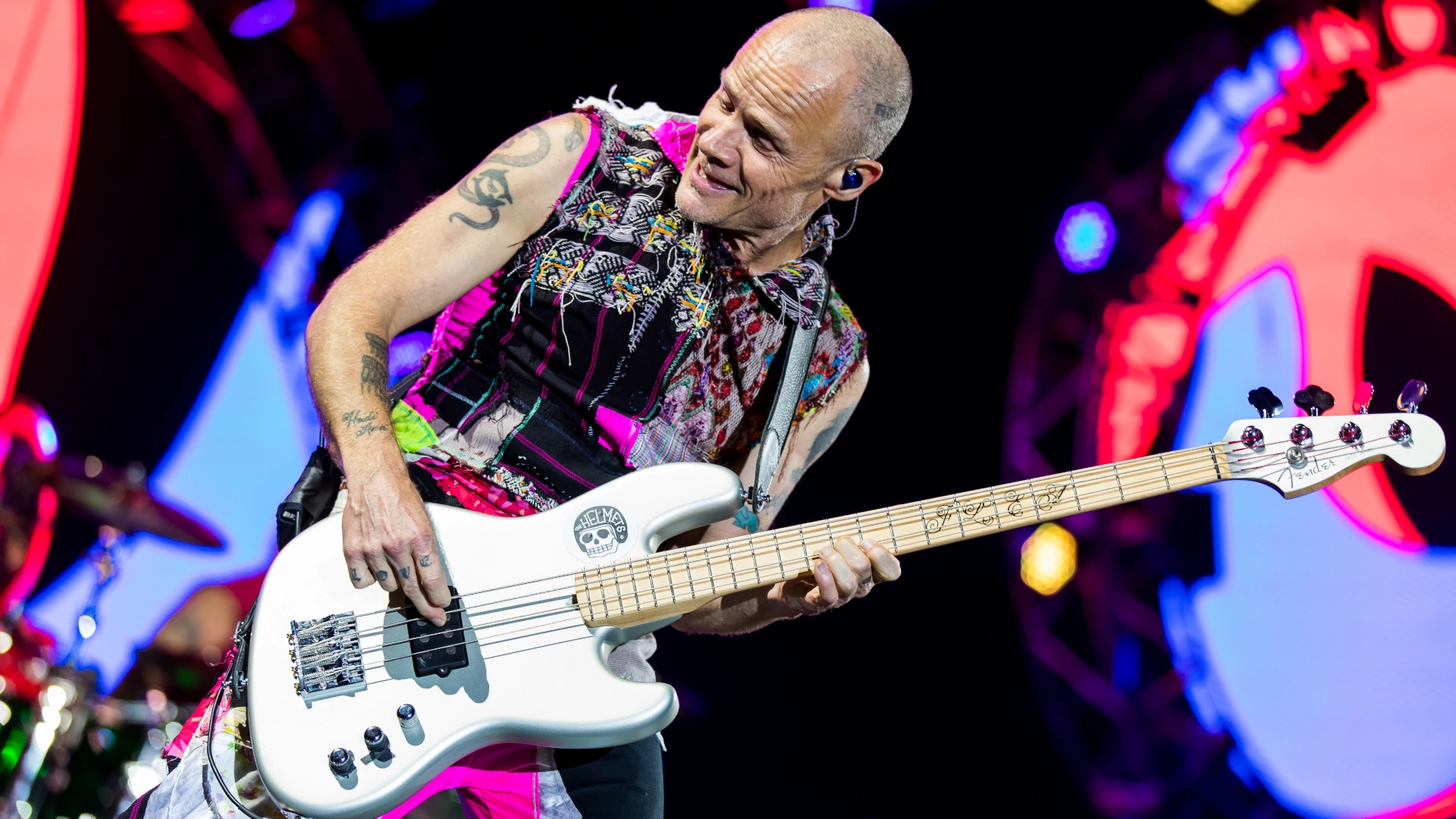 Flea of Red Hot Chili Peppers playing a silver Fender Jazz Bass on stage with colourful background.