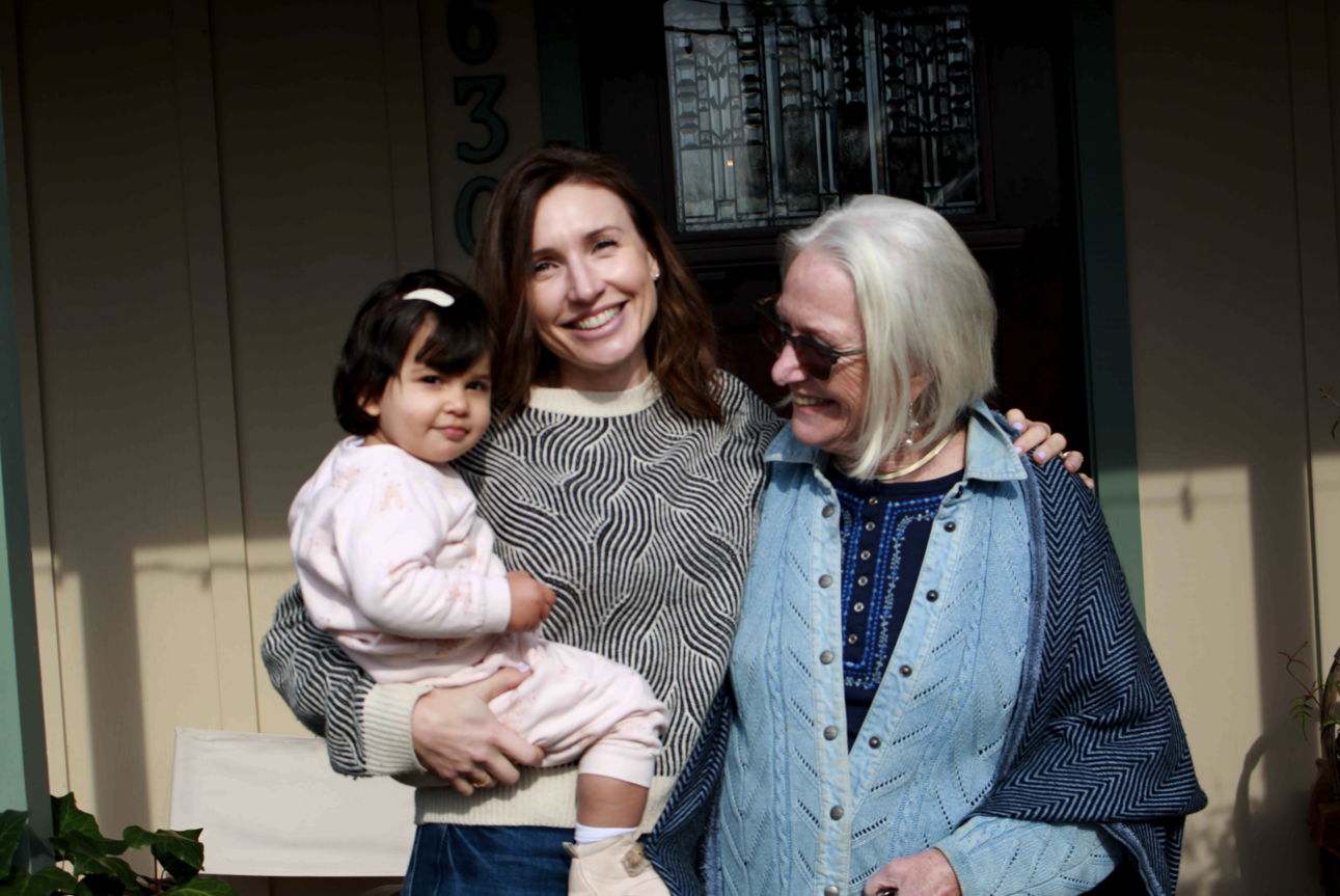 A woman holding a child stands beside an older woman, smiling outdoors.