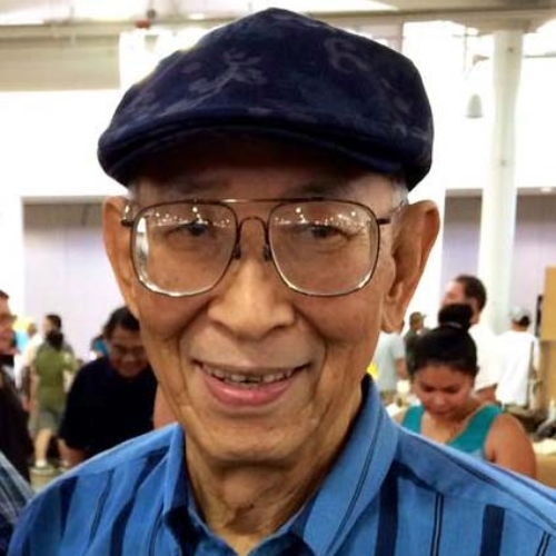 Elderly man smiling, wearing glasses and a blue cap, in a crowded setting.