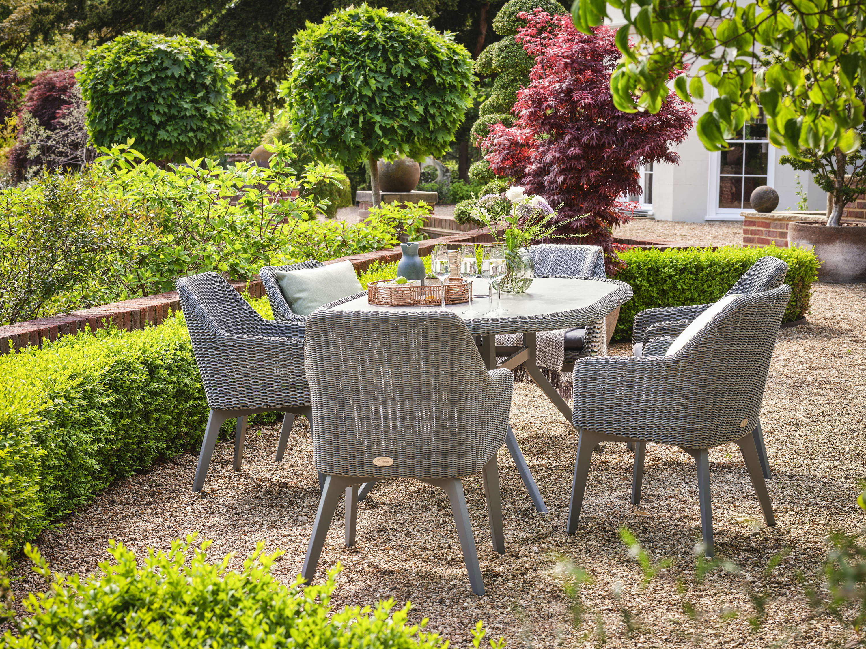 Grey garden rattan dining set with 6 dining armchairs and an oval table surrounded by hedges and trees.