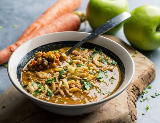 Bowl of soup with carrots and apples in the background.